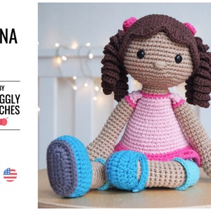 Pode incluir: Uma boneca de crochê com cabelo castanho, um vestido rosa e sapatos azuis. A boneca está sentada em uma superfície de madeira. A boneca se chama "Nina" e é de "Snuggly Stitches".