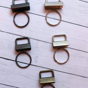 May include: Three sets of key rings with different finishes: black, silver, and antique brass. Each set includes a square-shaped key ring with a loop and a separate ring.