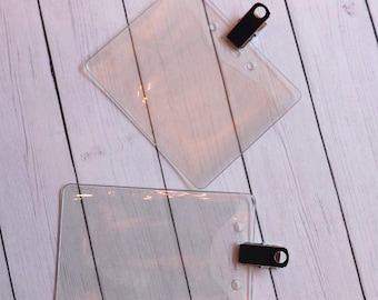 Puede incluir: Dos portagajetas de plástico transparente con clips negros. Los portagajetas son rectangulares y tienen esquinas redondeadas. Los clips están unidos a la parte superior de los portagajetas.