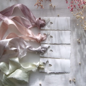 May include: Four rectangular white paper tags with deckled edges and a die-cut flower shape at the top. Each tag is tied with a long, flowing silk ribbon in varying pastel shades of dusty mauve, soft pink, light grey, and pale sage green.