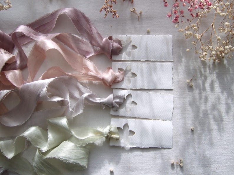 May include: Four rectangular white paper tags with deckled edges and a die-cut flower shape at the top. Each tag is tied with a long, flowing silk ribbon in varying pastel shades of dusty mauve, soft pink, light grey, and pale sage green.