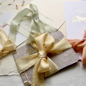 May include: A collection of stationery items including a small bound booklet with a textured gray cover and a gold ribbon bow, alongside white cards featuring gold foil text and accents, each tied with frayed silk ribbons in sage, cream, and peach.