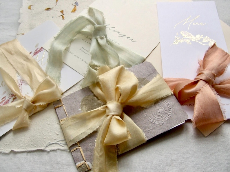 May include: A collection of stationery items including a small bound booklet with a textured gray cover and a gold ribbon bow, alongside white cards featuring gold foil text and accents, each tied with frayed silk ribbons in sage, cream, and peach.