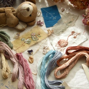 May include: A collection of embroidery floss in various colors, including green, pink, blue, and brown, arranged on a white fabric surface. There are also several small pieces of fabric with embroidered designs, including a floral pattern and a leafy branch. A small brown teddy bear is lying on the fabric.