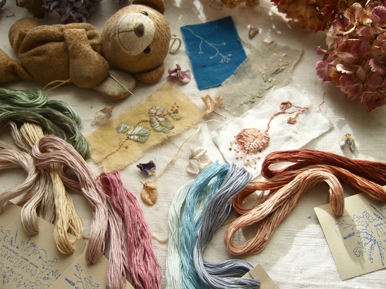 May include: A collection of embroidery floss in various colors, including green, pink, blue, and brown, arranged on a white fabric surface. There are also several small pieces of fabric with embroidered designs, including a floral pattern and a leafy branch. A small brown teddy bear is lying on the fabric.
