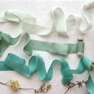 May include: Assortment of green and white silk ribbon spools. The ribbon is arranged in a decorative display on a white background.