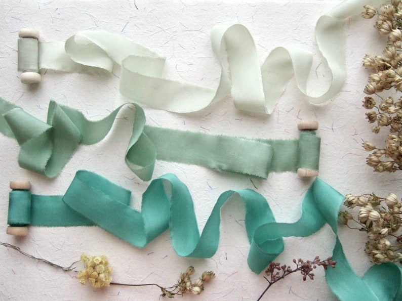May include: Assortment of green and white silk ribbon spools. The ribbon is arranged in a decorative display on a white background.