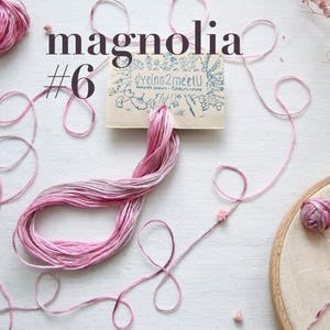 Vue aérienne de fil à broder rose et blanc bariolé, intitulé "magnolia #6". L'image comprend une pelote de fil, des fils lâches et un cercle à broder avec des broderies florales. Une petite carte avec "Dyeing2meetU" est également présente.