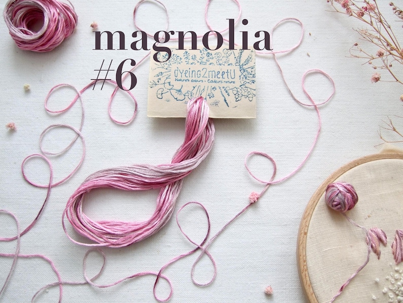 Vue aérienne de fil à broder rose et blanc bariolé, intitulé "magnolia #6". L'image comprend une pelote de fil, des fils lâches et un cercle à broder avec des broderies florales. Une petite carte avec "Dyeing2meetU" est également présente.