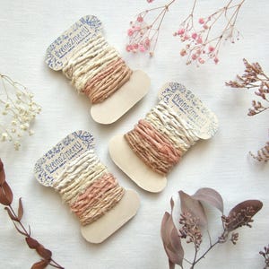 May include: Three spools of ribbon wrapped in cream and dusty rose colored fabric strips. Each spool features a cardboard tag printed with the text dyeing2meetU. The ribbons have a textured, frayed appearance and are wound around flat, light-colored cardboard bobbins.