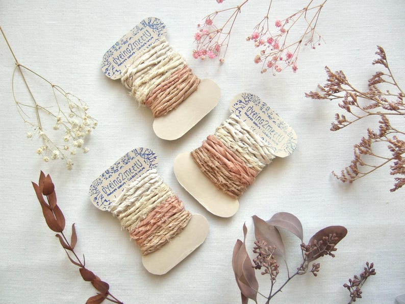 May include: Three spools of ribbon wrapped in cream and dusty rose colored fabric strips. Each spool features a cardboard tag printed with the text dyeing2meetU. The ribbons have a textured, frayed appearance and are wound around flat, light-colored cardboard bobbins.