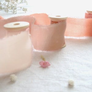 Peut inclure: Deux bobines de ruban de soie rose pâle aux bords effilochés, sur un fond de lin blanc. Le ruban est enroulé autour de bobines en bois.