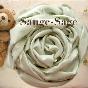 May include: A sage green silk scarf is arranged in a rose shape. The scarf is soft and flowing, with a subtle sheen. The text "Sauge-Sage" is printed on the scarf.