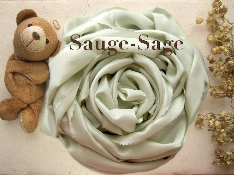 May include: A sage green silk scarf is arranged in a rose shape. The scarf is soft and flowing, with a subtle sheen. The text "Sauge-Sage" is printed on the scarf.