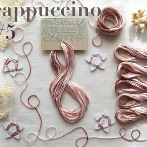 Fils à broder couleur cappuccino, composé de fils beige, rose et bruns. L'image présente des bobines, des écheveaux et des brins de fil lâches, ainsi que des décorations en forme d'étoile et une petite carte avec "Dyeing2meetU"