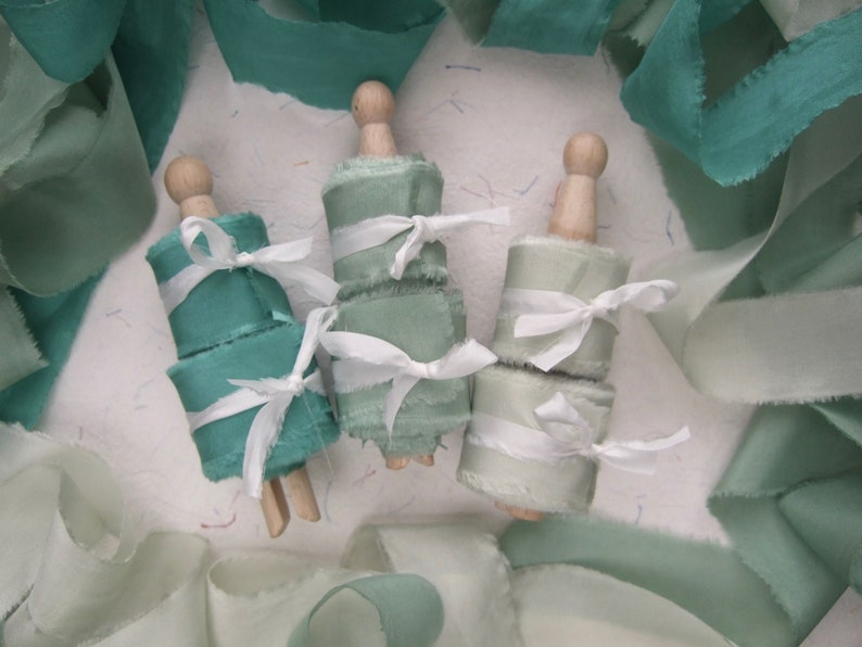 May include: Three spools of teal and green silk ribbon with frayed edges, tied with white ribbon and wooden spools.