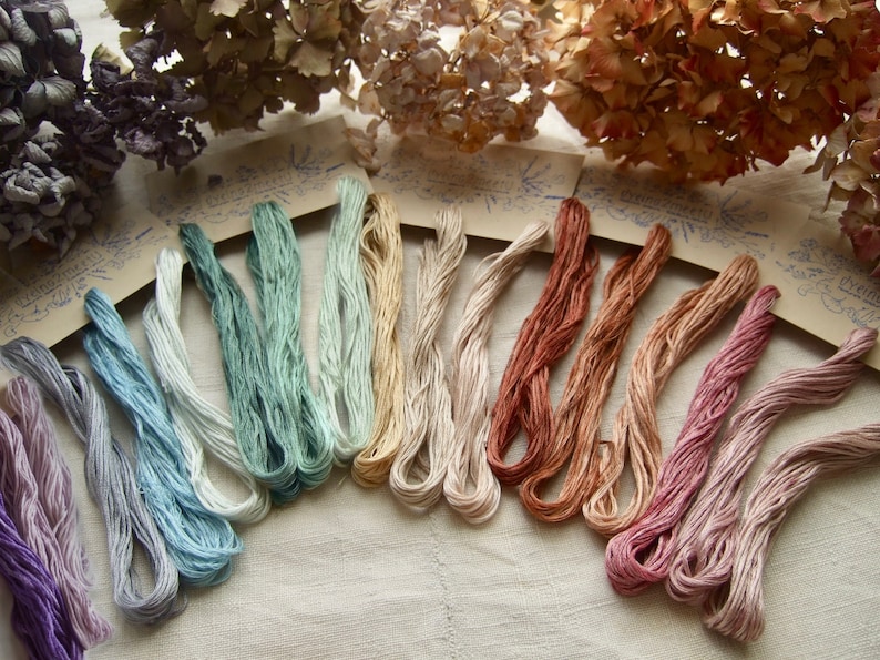 Une collection de fil à broder de coton à 6 brins, dans un arc-en-ciel de couleurs: nuances de bleu, vert, jaune, orange, violet, rose... Le fil est disposé en éventail sur un fond de tissu blanc. Les fils sont teints à la main en France par Idy de Dyeing2meetU