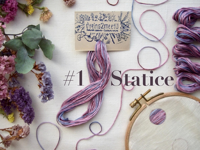 Un cercle à broder avec le texte "#1 Statice" et une échevette de fil rose, parme et bleu. Plusieurs échevettes de fil à broder sont disposés sur du lin blanc, ainsi qu'une petite carte "Dyeing2meetU" et des fleurs séchées