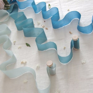 Peut inclure: Deux bobines de ruban dégradé dans des tons de bleu et de blanc. Le ruban est disposé en forme de vague sur une surface blanche.