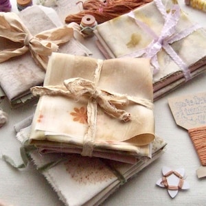 Puede incluir: Montones de papel hecho a mano atados con cintas y cordeles, con tintes naturales y estampados botánicos. Bobinas de hilo y suministros de artesanía están dispuestos alrededor del papel.