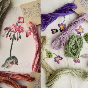 May include: Assortment of embroidery floss in various shades of green, pink, purple, and brown. The floss is arranged on a white fabric background with embroidered flowers and leaves.
