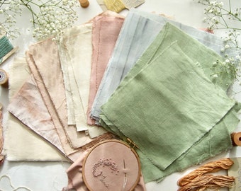 May include: A collection of square fabric swatches in shades of sage green, dusty rose, cream, and light gray, arranged in a fan shape. The fabric pieces feature raw, frayed edges. An embroidery hoop with pink fabric and a needle is positioned below.
