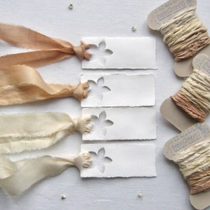 May include: Four rectangular white paper tags with a die-cut flower shape, each tied with a frayed silk ribbon in varying shades of beige and cream. Three cardboard spools are wrapped with matching hand-dyed silk ribbons, labeled dyeing2meetU.