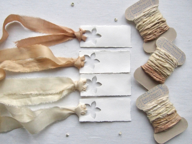May include: Four rectangular white paper tags with a die-cut flower shape, each tied with a frayed silk ribbon in varying shades of beige and cream. Three cardboard spools are wrapped with matching hand-dyed silk ribbons, labeled dyeing2meetU.