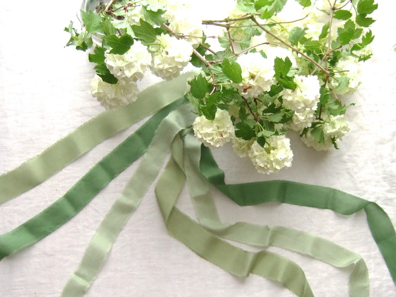 May include: Four green ombre ribbon spools with a bouquet of white flowers in the background.