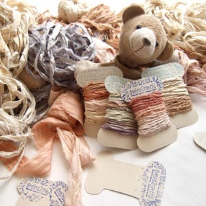 May include: A collection of hand-dyed silk ribbons in neutral, peach, and grey tones, some wound on cardboard spools labeled dyeing2meetU, alongside a small brown plush teddy bear and loose bundles of textured fabric strips in various shades.