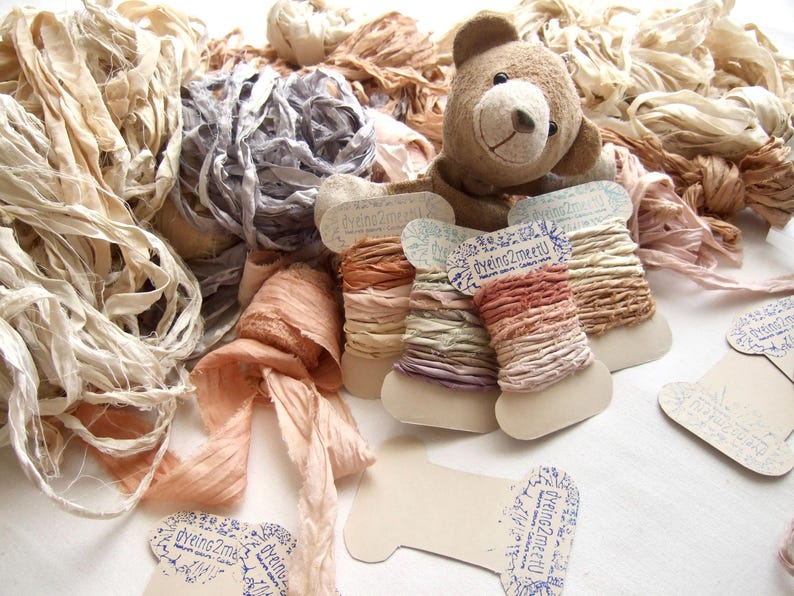 May include: A collection of hand-dyed silk ribbons in neutral, peach, and grey tones, some wound on cardboard spools labeled dyeing2meetU, alongside a small brown plush teddy bear and loose bundles of textured fabric strips in various shades.
