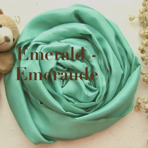 May include: A soft, emerald green fabric is folded into a rose shape. The fabric is smooth and silky. The text "Emerald - Emeraude" is written in brown text on the fabric.