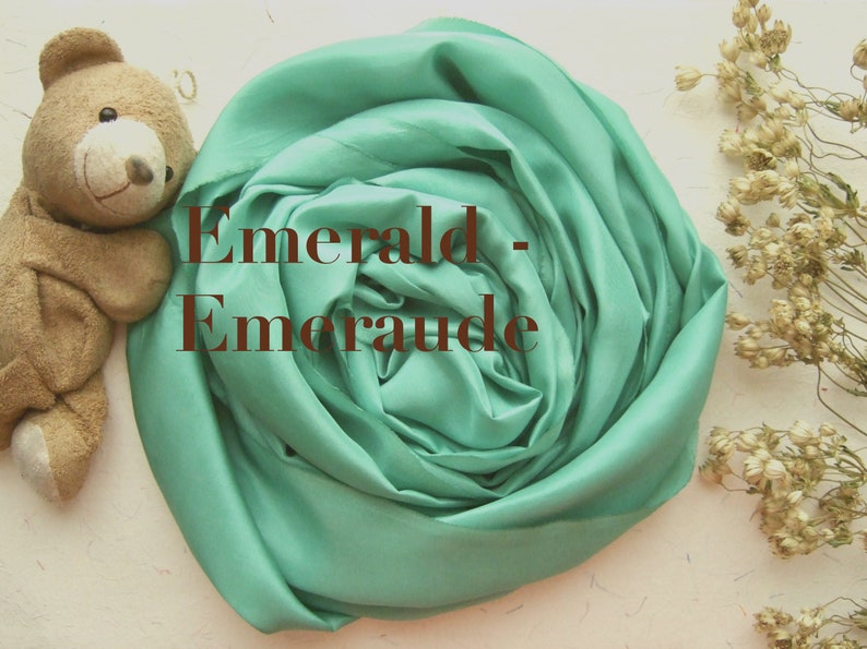May include: A soft, emerald green fabric is folded into a rose shape. The fabric is smooth and silky. The text "Emerald - Emeraude" is written in brown text on the fabric.
