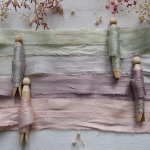 May include: Four strips of frayed-edge silk ribbon in shades of sage green, light gray, dusty mauve, and soft pink. Each ribbon is wrapped around a wooden clothespin, arranged horizontally across the frame with delicate dried flowers nearby.