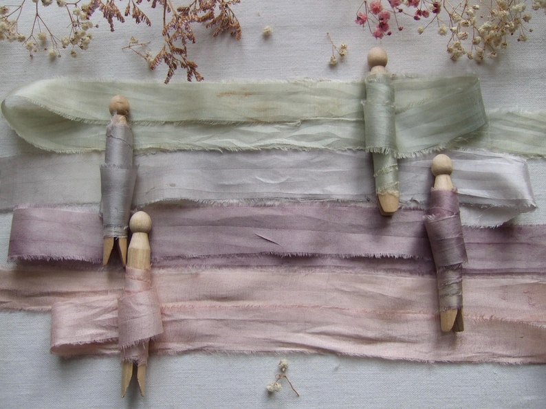 May include: Four strips of frayed-edge silk ribbon in shades of sage green, light gray, dusty mauve, and soft pink. Each ribbon is wrapped around a wooden clothespin, arranged horizontally across the frame with delicate dried flowers nearby.