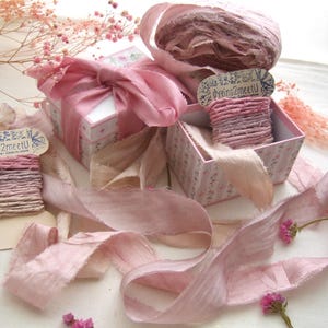 May include: A collection of dusty pink silk ribbons, some wound around cardboard spools labeled dyeing2meetU, alongside two small square gift boxes with floral patterns and pink ribbon bows, accented by sprigs of dried pink baby's breath flowers.