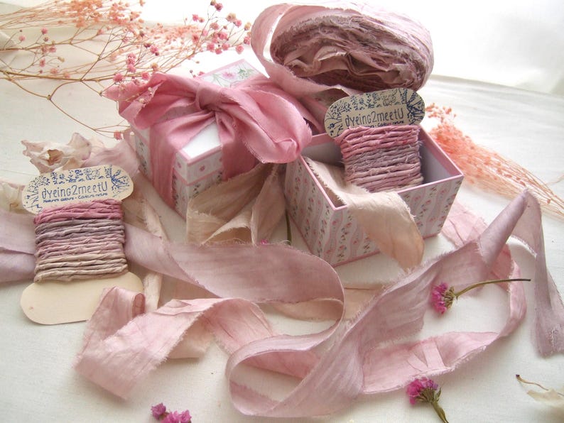 May include: A collection of dusty pink silk ribbons, some wound around cardboard spools labeled dyeing2meetU, alongside two small square gift boxes with floral patterns and pink ribbon bows, accented by sprigs of dried pink baby's breath flowers.