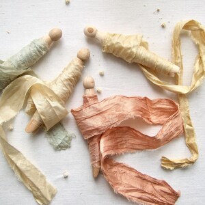 May include: Three wooden clothespins wrapped with frayed silk ribbons in shades of sage green, pale yellow, and dusty peach. Each clothespin features a rounded top, with the ribbons wound around the body and trailing off in loose, textured lengths.