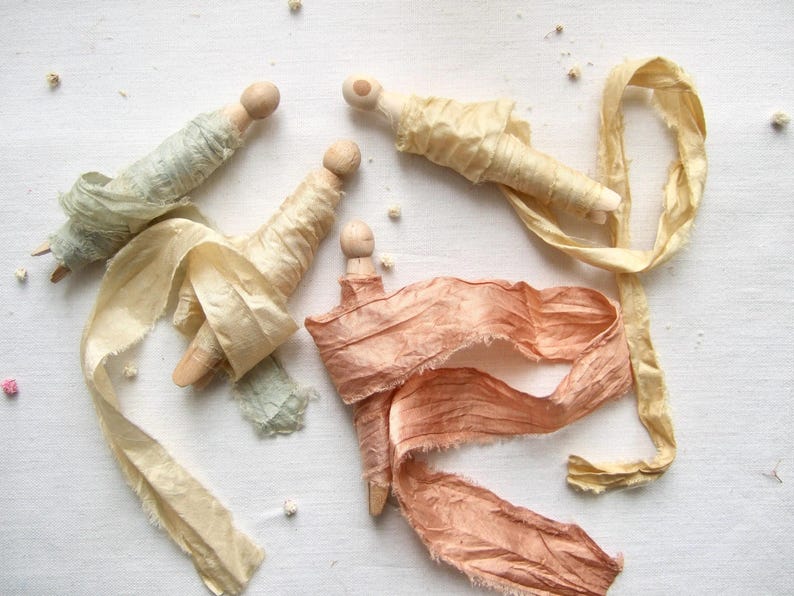 May include: Three wooden clothespins wrapped with frayed silk ribbons in shades of sage green, pale yellow, and dusty peach. Each clothespin features a rounded top, with the ribbons wound around the body and trailing off in loose, textured lengths.