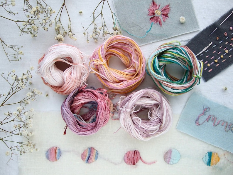 Plusieurs échevettes de fil à broder coloré dans des tons de rose, jaune et vert. L'image présente également des échantillons de tissu brodé avec des motifs floraux et abstraits, et le nom "Emma" brodé en cursive.