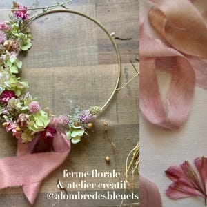 May include: A circular gold metal hoop adorned with dried flowers in shades of pink, green, and white, featuring a dusty pink silk ribbon tied at the bottom. The text "ferme florale & atelier cr&eacute;atif @alombredesbleuets" is printed at the bottom.