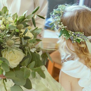 May include: A white bridal bouquet featuring white roses, green eucalyptus leaves, and small purple flowers. A flower crown with white and purple flowers and green leaves is worn by a girl.