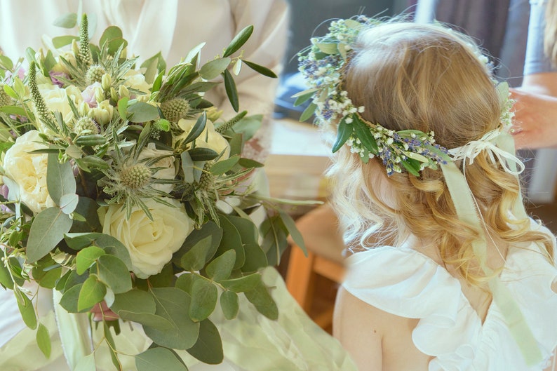 May include: A white bridal bouquet featuring white roses, green eucalyptus leaves, and small purple flowers. A flower crown with white and purple flowers and green leaves is worn by a girl.