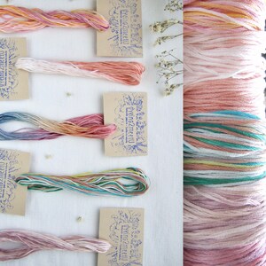 May include: Assortment of colorful embroidery floss skeins in shades of pink, orange, blue, and green, each with a small tag. A larger spool of floss is visible on the right. The tags have a blue stamp with text.