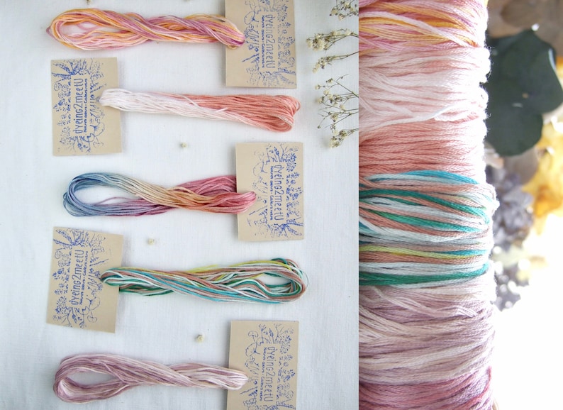 May include: Assortment of colorful embroidery floss skeins in shades of pink, orange, blue, and green, each with a small tag. A larger spool of floss is visible on the right. The tags have a blue stamp with text.