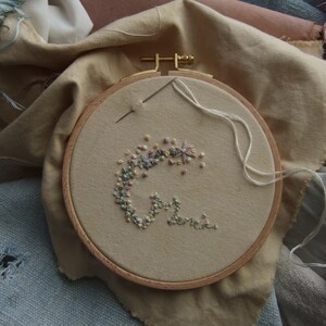 Un cercle à broder avec un tissu beige orné d'un motif floral brodé avec du fil de couleur pastel. Le cercle en bois est posé sur un fond de divers tissus et de bobines de fil, suggérant un projet artisanal.