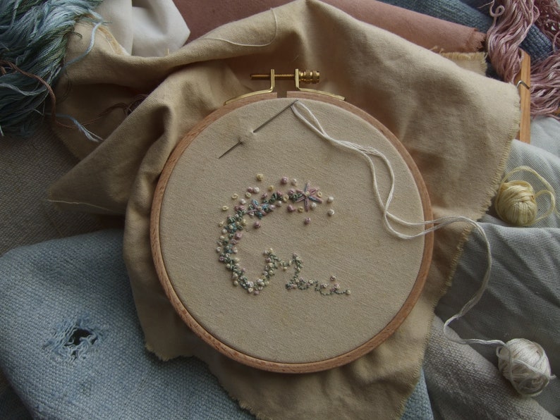 Un cercle à broder avec un tissu beige orné d'un motif floral brodé avec du fil de couleur pastel. Le cercle en bois est posé sur un fond de divers tissus et de bobines de fil, suggérant un projet artisanal.