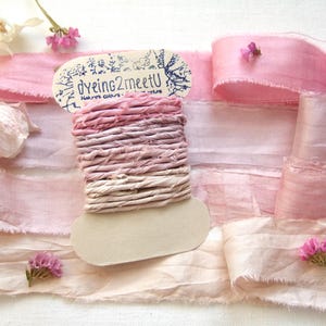 May include: A spool of ombre pink hand-dyed silk ribbon with frayed edges, featuring the text "dyeing2meetU" on the cardboard label. The spool is surrounded by lengths of matching light pink silk ribbon and small clusters of pink flowers.