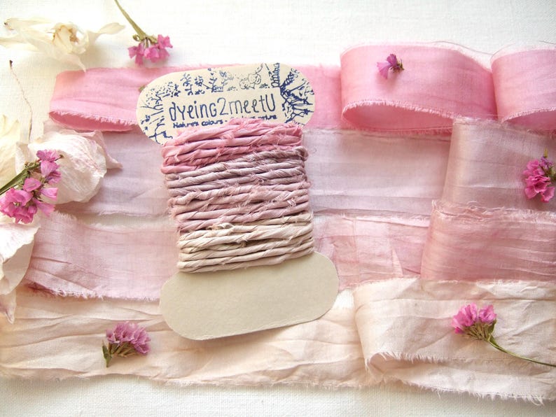 May include: A spool of ombre pink hand-dyed silk ribbon with frayed edges, featuring the text "dyeing2meetU" on the cardboard label. The spool is surrounded by lengths of matching light pink silk ribbon and small clusters of pink flowers.