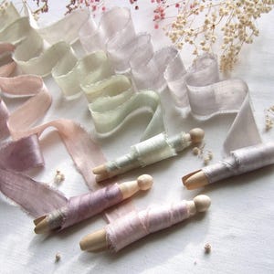 May include: A collection of frayed-edge silk ribbons in soft pastel shades of mauve, dusty pink, sage green, and light gray. Several ribbons are neatly wound around wooden clothespins, while others are draped in loose, flowing waves.